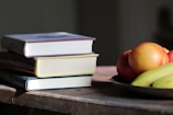 A stack of three books with colorful spines sits on a wooden table, next to a metal plate containing apples and bananas. The scene is illuminated by natural light, casting soft shadows.