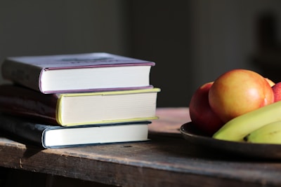 A stack of three books with colorful spines sits on a wooden table, next to a metal plate containing apples and bananas. The scene is illuminated by natural light, casting soft shadows.