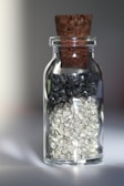 A small glass jar with a cork stopper, containing two distinct layers of crystalline materials. The upper layer consists of dark, shiny crystals, while the lower layer contains lighter, silver-like crystals.