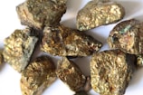 A collection of rough, metallic-looking rocks with a golden sheen, possibly pyrite, are scattered on a white surface, reflecting light.