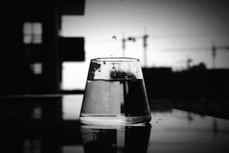 A clear glass of water sits upside down on a reflective surface, with raindrops visible on its exterior. The background is blurred, featuring an urban skyline with cranes and buildings, indicating a possible construction site. The image has a black and white color scheme, providing a contrast between the clear glass and the dark outlines of the background.