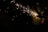 Sparks fly from a handheld power tool operated by a person, illuminating parts of the scene against a dark background. The person appears in partial silhouette, with the bright sparks creating a stark contrast.