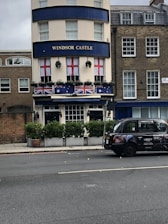 A building with a sign labeled 'Windsor Castle' on its facade. Red and white flags with a red cross pattern hang from the windows, and flags with blue and red elements are displayed on the balcony below. Potted plants with greenery are placed in front of the building. A black taxi with advertisements is parked on the street next to the curb. The surrounding architecture has a traditional and classic style.