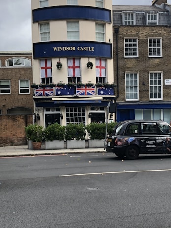 A building with a sign labeled 'Windsor Castle' on its facade. Red and white flags with a red cross pattern hang from the windows, and flags with blue and red elements are displayed on the balcony below. Potted plants with greenery are placed in front of the building. A black taxi with advertisements is parked on the street next to the curb. The surrounding architecture has a traditional and classic style.