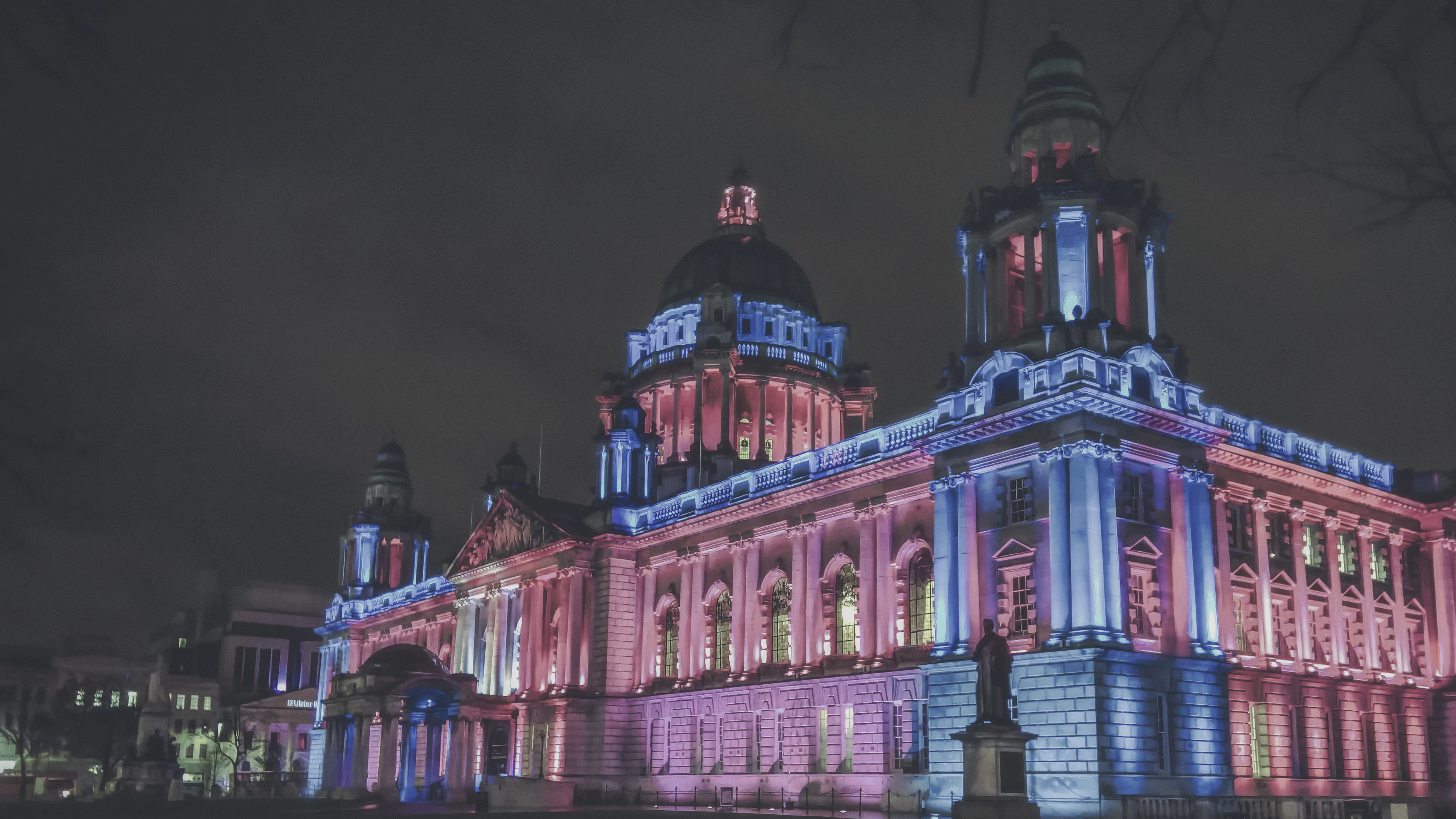 Belfast City Hall illuminated in pink and blue light at night, its dome glowing above a statue in the foreground.