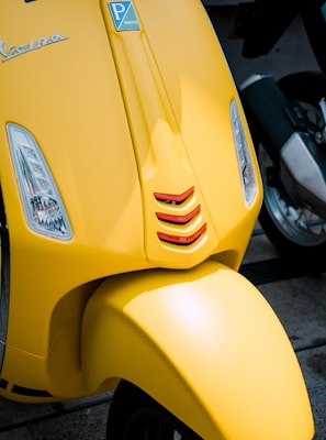 A close-up of the front of a yellow Vespa scooter, featuring a Piaggio emblem, two small headlights integrated into the body, and red accents near the wheel cover.