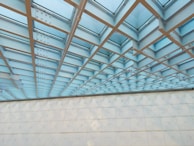 A geometric ceiling structure with a grid pattern dominates the scene. The ceiling consists of intersecting steel beams with panels of glass in between, allowing natural light to filter through. The wall below is made up of smooth, cream-colored panels lined with a series of small circular fixtures.