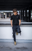 A young person with tousled hair stands casually against a concrete railing. They are dressed in a black t-shirt, dark pants, and high-top sneakers in shades of brown and black. The background is an urban setting with a parking structure and some blurred elements suggesting movement.