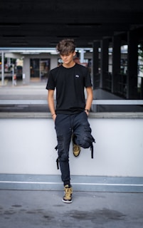 A young person with tousled hair stands casually against a concrete railing. They are dressed in a black t-shirt, dark pants, and high-top sneakers in shades of brown and black. The background is an urban setting with a parking structure and some blurred elements suggesting movement.
