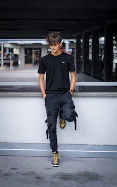 A young person with tousled hair stands casually against a concrete railing. They are dressed in a black t-shirt, dark pants, and high-top sneakers in shades of brown and black. The background is an urban setting with a parking structure and some blurred elements suggesting movement.