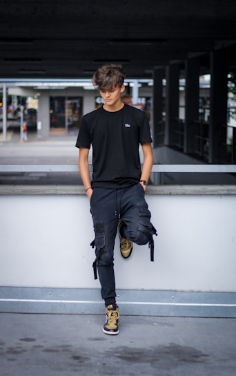 A young person with tousled hair stands casually against a concrete railing. They are dressed in a black t-shirt, dark pants, and high-top sneakers in shades of brown and black. The background is an urban setting with a parking structure and some blurred elements suggesting movement.