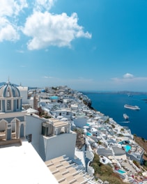 A picturesque coastal town with whitewashed buildings and blue domes perched on cliffs overlooking a vibrant blue sea. A large cruise ship is visible in the distance, accompanied by smaller boats. The sky is clear with a few fluffy clouds, adding to the serene ambiance.