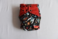 A colorful diaper with a space-themed pattern featuring rockets and stars, accented with red and black snap buttons on the front. The background is predominantly black with vibrant illustrations.