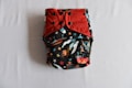 A colorful diaper with a space-themed pattern featuring rockets and stars, accented with red and black snap buttons on the front. The background is predominantly black with vibrant illustrations.