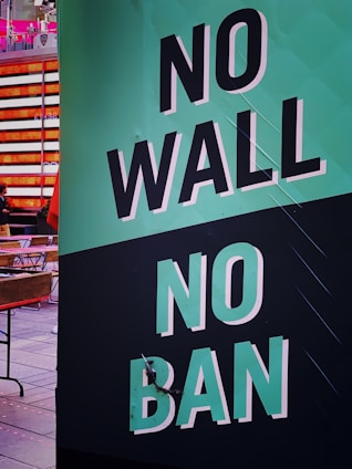 A large sign displays the message 'NO WALL NO BAN' in bold, white letters on a green and black background. The setting appears to be an urban environment with buildings and people visible in the background.