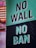 A large sign displays the message 'NO WALL NO BAN' in bold, white letters on a green and black background. The setting appears to be an urban environment with buildings and people visible in the background.