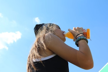 A person is drinking from an orange bottle on a clear, sunny day. The individual is wearing a black tank top and a headband, with bracelets on their wrist. The sky is bright blue with a few small clouds.