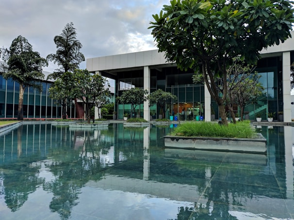 A modern building with large glass windows is surrounded by lush green trees and a reflective water feature. The trees are planted in geometric concrete beds along the edge of a calm pool, creating a serene and harmonious environment. The sky is partly cloudy, adding to the tranquil atmosphere.