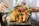 A culinary presentation featuring fried samosas, prawns, and an assortment of fresh vegetables and berries on a dark serving board. The background shows blurred figures handling a dish, adding a dynamic element to the composition.