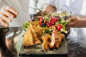 A culinary presentation featuring fried samosas, prawns, and an assortment of fresh vegetables and berries on a dark serving board. The background shows blurred figures handling a dish, adding a dynamic element to the composition.