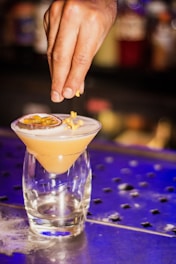 A hand decorates a cocktail with garnish, featuring a drink in an elegant glass with a frothy top and a slice of passion fruit. The background is dimly lit, with blurred bottles providing an intimate bar setting.