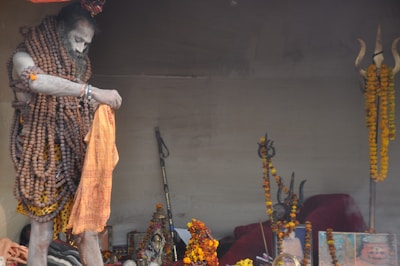 A person is adorned with numerous rudraksha beads and covered in ash, holding an orange cloth. The setting includes various religious artifacts, including a trident embellished with marigold garlands, and religious images or idols.