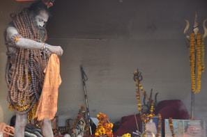 A person is adorned with numerous rudraksha beads and covered in ash, holding an orange cloth. The setting includes various religious artifacts, including a trident embellished with marigold garlands, and religious images or idols.