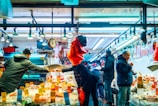 A vibrant market scene with several people engaged in buying and selling seafood under bright overhead lights. The counter is lined with various seafood items, some of which have price tags displayed prominently. A scale is being used by one person, while others interact or examine the goods. The atmosphere is bustling and lively.