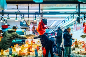 A vibrant market scene with several people engaged in buying and selling seafood under bright overhead lights. The counter is lined with various seafood items, some of which have price tags displayed prominently. A scale is being used by one person, while others interact or examine the goods. The atmosphere is bustling and lively.
