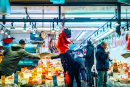 A vibrant market scene with several people engaged in buying and selling seafood under bright overhead lights. The counter is lined with various seafood items, some of which have price tags displayed prominently. A scale is being used by one person, while others interact or examine the goods. The atmosphere is bustling and lively.