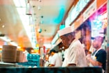 A bustling diner kitchen with two individuals dressed in white uniforms, one wearing a white hat, focused on their tasks. Stacks of plates are visible in the foreground, and colorful neon signs illuminate the background, creating a lively atmosphere.