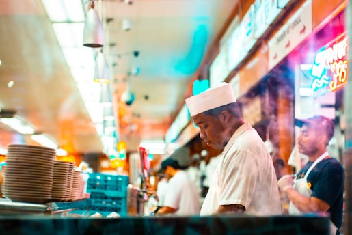 A bustling diner kitchen with two individuals dressed in white uniforms, one wearing a white hat, focused on their tasks. Stacks of plates are visible in the foreground, and colorful neon signs illuminate the background, creating a lively atmosphere.