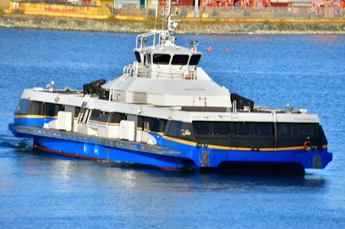 A modern ferry with a sleek design is cruising on calm blue water. The vessel is painted in shades of blue and white, with a yellow stripe near the waterline. The architecture of the ferry suggests a focus on functionality and passenger capacity. In the background, there is a glimpse of an industrial area with containers and machinery.
