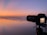 A DSLR camera is set on a tripod capturing a serene ocean view during sunset. The sky is a gradient of soft oranges and pinks reflecting on the wet sand. Gentle waves are visible approaching the shore.