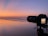 A DSLR camera is set on a tripod capturing a serene ocean view during sunset. The sky is a gradient of soft oranges and pinks reflecting on the wet sand. Gentle waves are visible approaching the shore.