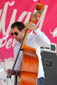 A man wearing sunglasses plays a double bass. He is dressed in a white shirt and the instrument is made of a rich, polished wood. In the background, a vibrant pink banner with white text is partially visible.