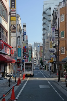 A bustling urban street scene featuring tall buildings with numerous colorful signs in various languages. The street is lined with bicycles and orange traffic cones, and a small white truck is parked along the road. The atmosphere suggests a busy commercial area.