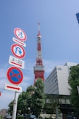 A prominent red and white communications tower stands tall against a clear blue sky. In the foreground, multiple road signs are visible, indicating various traffic regulations. The base of the tower is surrounded by greenery and a modern office building labeled 'Tata Consultancy Services'. The overall scene is urban with clear visibility.