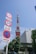 A prominent red and white communications tower stands tall against a clear blue sky. In the foreground, multiple road signs are visible, indicating various traffic regulations. The base of the tower is surrounded by greenery and a modern office building labeled 'Tata Consultancy Services'. The overall scene is urban with clear visibility.