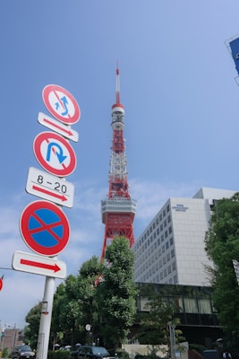 A prominent red and white communications tower stands tall against a clear blue sky. In the foreground, multiple road signs are visible, indicating various traffic regulations. The base of the tower is surrounded by greenery and a modern office building labeled 'Tata Consultancy Services'. The overall scene is urban with clear visibility.