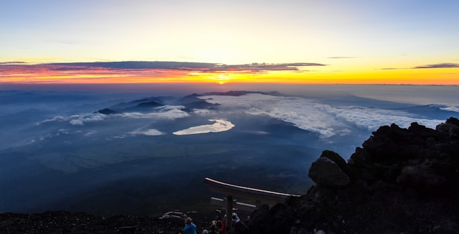 A breathtaking sunrise view from a mountaintop, showcasing a vibrant sky with hues of orange, pink, and yellow. The clouds float below the silhouetted mountain peaks and a serene body of water reflects the early morning light. Dark rocky terrain surrounds the foreground.