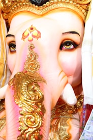 The image features a close-up of a Ganesha idol, showcasing intricate detailing with gold decorations on the trunk. The eyes are prominently expressive, outlined with dark makeup, and the forehead displays vibrant patterns. The vibrant colors and embellishments contribute to a sense of richness and reverence.
