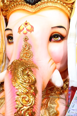 The image features a close-up of a Ganesha idol, showcasing intricate detailing with gold decorations on the trunk. The eyes are prominently expressive, outlined with dark makeup, and the forehead displays vibrant patterns. The vibrant colors and embellishments contribute to a sense of richness and reverence.