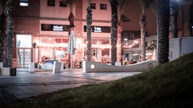 A small urban plaza features several palm trees and modern concrete benches. Tables and chairs are set up outside a brightly lit caf&eacute;, with string lights creating a warm atmosphere. The building fa&ccedil;ade includes signage, and the area is lightly populated, suggesting a calm evening setting.