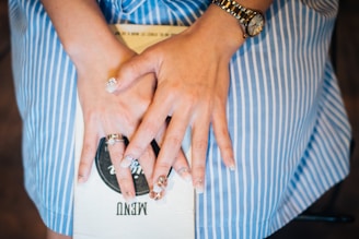 A person is wearing a striped blue and white dress, sitting with hands resting on a menu. The hands display multiple rings and manicured nails. A watch adorns one wrist, and the setting suggests a casual or dining environment.