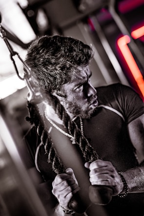 A man appears to be engaging in a strenuous workout, using a cable machine. His expression is focused and intense, suggesting effort and concentration. He is gripping the ends of two ropes and wearing a dark, tight-fitting athletic shirt, along with a necklace and a wrist accessory. The image captures the essence of determination in a gym environment.