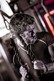 A man appears to be engaging in a strenuous workout, using a cable machine. His expression is focused and intense, suggesting effort and concentration. He is gripping the ends of two ropes and wearing a dark, tight-fitting athletic shirt, along with a necklace and a wrist accessory. The image captures the essence of determination in a gym environment.