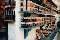 Shelves filled with a variety of jars containing preserves and sauces, labeled and neatly arranged. The jars are topped with brown paper and tied with string. There are different colors and types of preserves, creating a rustic and artisanal market display.