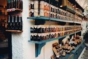 Shelves filled with a variety of jars containing preserves and sauces, labeled and neatly arranged. The jars are topped with brown paper and tied with string. There are different colors and types of preserves, creating a rustic and artisanal market display.