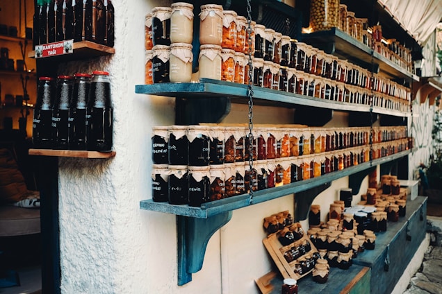 Shelves filled with a variety of jars containing preserves and sauces, labeled and neatly arranged. The jars are topped with brown paper and tied with string. There are different colors and types of preserves, creating a rustic and artisanal market display.